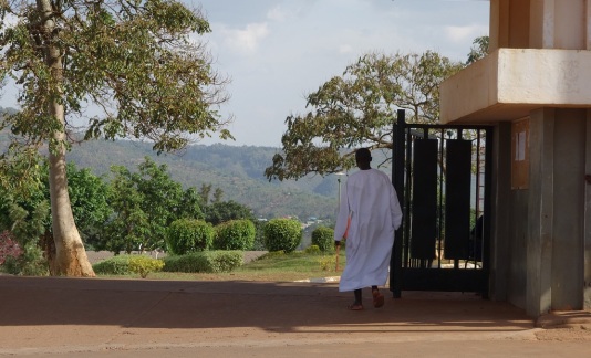A man wearing a white Muslim dress enters a mosque's yard. The background of the picture shows the hilly Rwandan landscape.