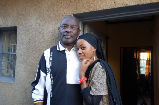 Yahaya Nesengiyumva is standing in front of the door of his house. Next to him one of his daughters.